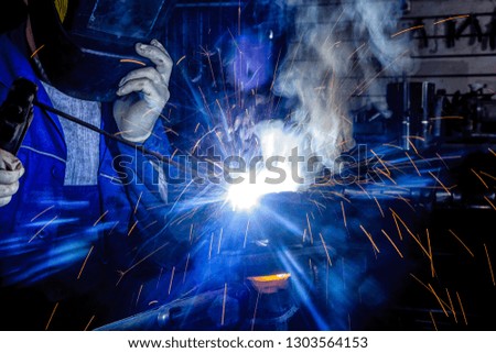 Man welder in welding mask, building uniform and protective gloves welds metal car part with welding machine in auto repair shop, in the background construction site