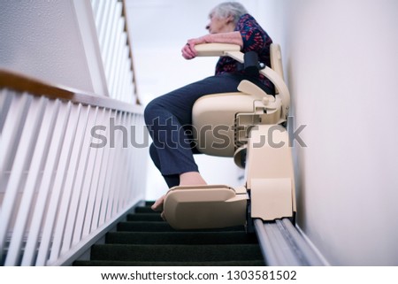 Senior Woman Sitting On Stair Lift At Home To Help Mobility Royalty-Free Stock Photo #1303581502