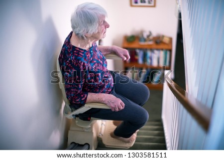 Senior Woman Sitting On Stair Lift At Home To Help Mobility Royalty-Free Stock Photo #1303581511