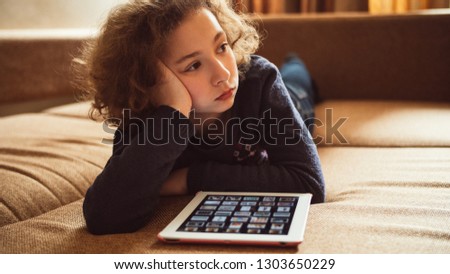 Bored little girl looking photos and playing computer games on the tablet. Child entertainment, leisure and education with help of modern technologies. Mobile apps for kids.
