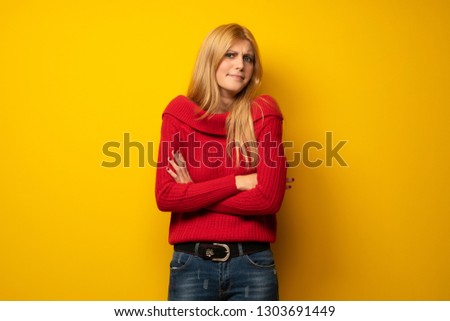 Blonde woman over yellow wall making doubts gesture while lifting the shoulders