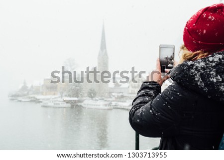 Woman in red hat taking picture of Hallstatt old town during snowfall, Austria.
