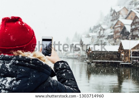 Woman in red hat taking picture of lakefront Hallstatt old town during snowfall, Austria.
