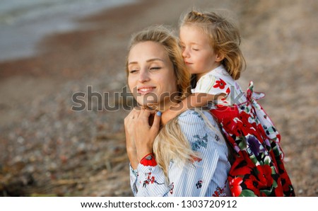 Happy and friendly family at sea. Emotional and positive little girl hugs her beautiful and young mother for neck against the beach. Childhood. Summertime. Summer vacation. Lifestyle. Positive energy