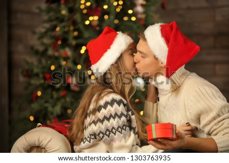 Loving young couple in Santa hats and with Christmas gift kissing on sofa at home