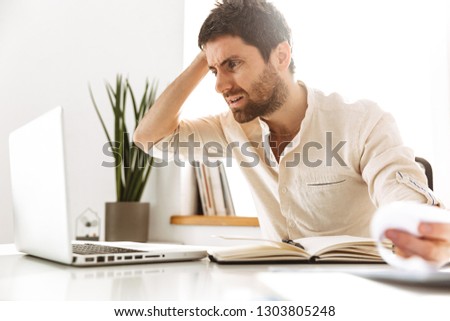 Portrait of uptight businessman 30s wearing white shirt working with laptop and paper documents while sitting in bright office Royalty-Free Stock Photo #1303805248