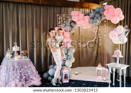 Happy family: mother and son. Portrait of young parent keep in arms, hugging child kid boy, celebrating birthday holiday party on pink, grey background with air balloons.