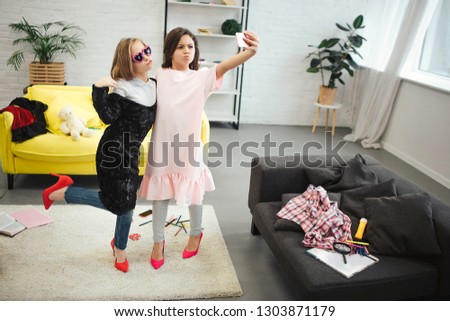 Stylish young teenagers stand in room and pose on phone camera. They wear clothes for adult women. Brunette hold camera. They take selfie.