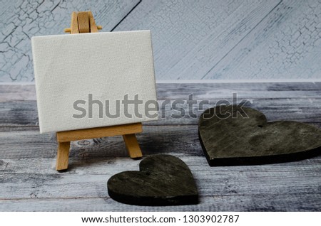 Small easel with a blank canvas over white and two dark wooden hearts. Old wooden background and big copy space for your sign and text