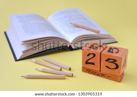 Perpetual wooden calendar with date of February 23, an opened book and pencils on a yellow background with copy space. Concept of Defender of the Fatherland Day, courage, strength. Horizontal photo