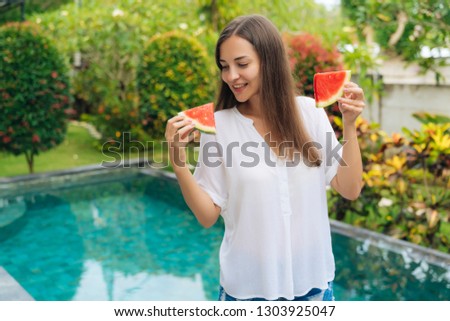 Portrait of pretty happy girl with delight holding watermelon pieces in her hands. Attractive brunette female eating fruits near swimming pool. Concept fresh, healthy food, vegetarian