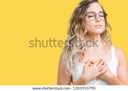 Beautiful young blonde woman wearing glasses over isolated background smiling with hands on chest with closed eyes and grateful gesture on face. Health concept.