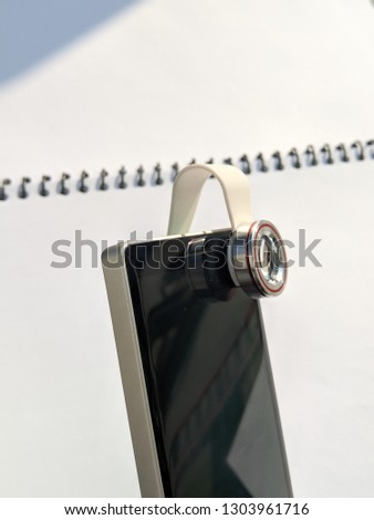Smartphone lens installed on front camera of smart phone, mobile phone with white isolated background. Lens clip mounted on mobile phone camera. Macro, Fisheye, wide angle lens for smartphones.
