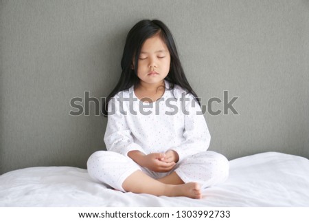 Asian child cute or kid girl sit meditation with peace or quiet and relax for mindfulness and concentrated breath on wake up in morning on bedroom at home and wear white dress on gray background