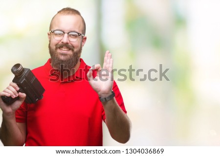 Young caucasian hipster man filmming using retro video camera over isolated background doing ok sign with fingers, excellent symbol