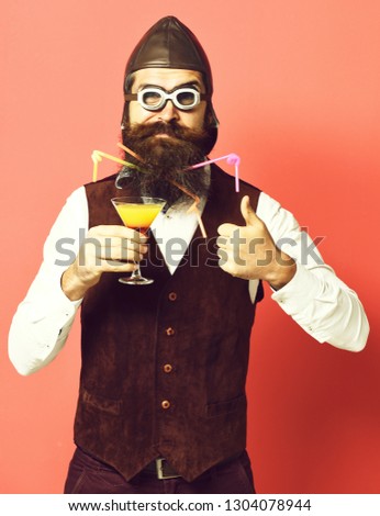 handsome bearded pilot or aviator man with long beard and mustache on serious face holding glass of alcoholic beverage in vintage suede leather waistcoat with hat and glasses on red studio background.