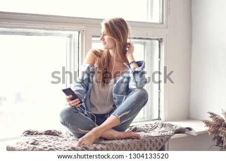young beautiful woman sits on a windowsill and listens to a podcast or radio on her mobile phone