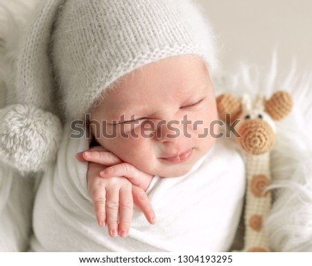 Little baby in hat covered in white blanket sleeping in basket with toy