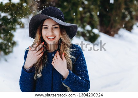 Beautiful young woman smile in the winter forest. snowfall. Blue coat and hat. Nice orange nails. Red lips. Lush blond hair. Winter holiday concept.
