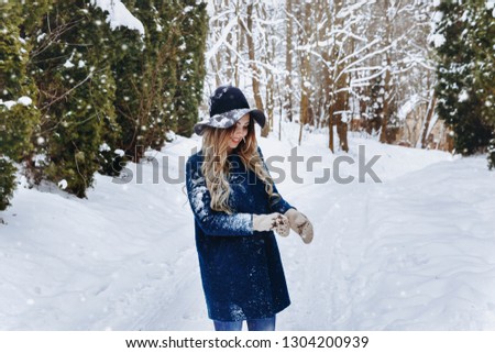 srtylish happy young woman walk in winter forest. Wear knitted gloves, blue coat and black hat. Snowfall. Christmas, winter holidays concept.
