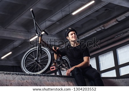 Young Bmx rider relaxing after practicing tricks with his bike in a skatepark indoors