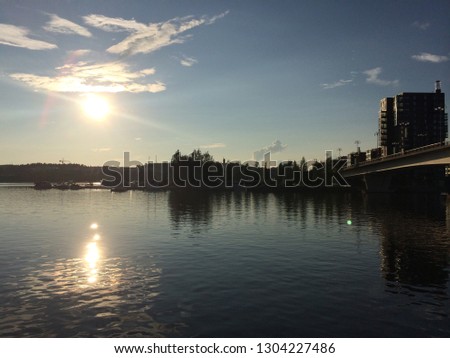 Picture of beautiful sun over water or a lake in Finland during the summer. Includes a bridge, boats, and a building.