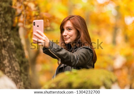 A young woman with reddish hair is taking selfies with her mobile phone in an autumnal forest of yellow and ocher colors in Montanchez, Caceres, Extremadura