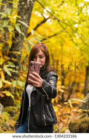 A young woman with reddish hair is taking selfies with her mobile phone in an autumnal forest of yellow and ocher colors in Montanchez, Caceres, Extremadura
