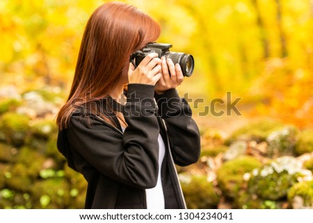 A young woman with reddish hair takes pictures with a reflex camera in an autumnal forest of yellow and ocher colors in Montanchez, Caceres, Extremadura