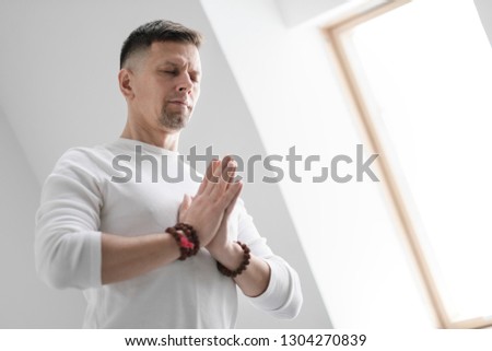 Male brunette with a beard in white clothes on a white background with his hands folded on his chest. Meditation and prayer, yoga.