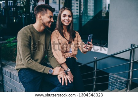 Positive woman looking at camera while holding mobile phone and taking photo with boyfriend outdoors,romantic couple feeling happiness together on free time using smartphone camera making selfie