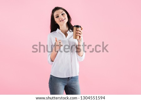 attractive brunette girl holding coffee to go and showing thumb up, isolated on pink