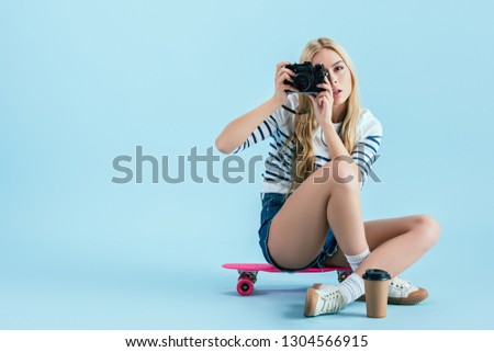 Studio shot of girl sitting on longboard and taking picture on blue background