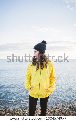 Portrait of a young brunette woman on the beach wearing a raincoat and hat.