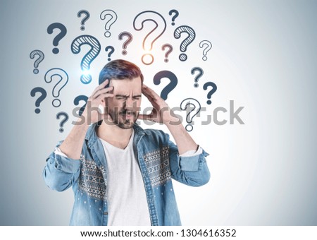 Portrait of bearded young man wearing jeans shirt and having strong headache standing near gray wall with question marks drawn on it. Mock up