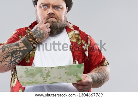 Isolated on the grey background photo of bearded fat traveler in red Hawaiian shirt thoughtfully touching his beard while standing with a map