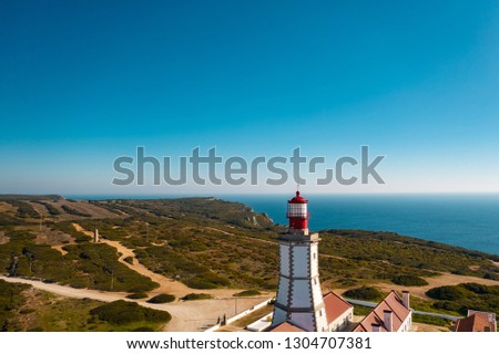 Aerial; drone view of white-red lighthouse Cabo Espichel on the edge of the earth; turquoise water of Atlantic ocean stretching to horizon; beautiful portuguese viewpoint with old historic sightseeing