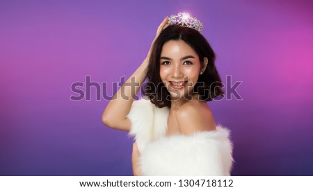 Portrait of Miss Pageant Beauty Contest in white fur Evening dress light Diamond Crown, LGBT transgender Asian Woman fashion make up black hair style, studio lighting gradient shade purple background Royalty-Free Stock Photo #1304718112