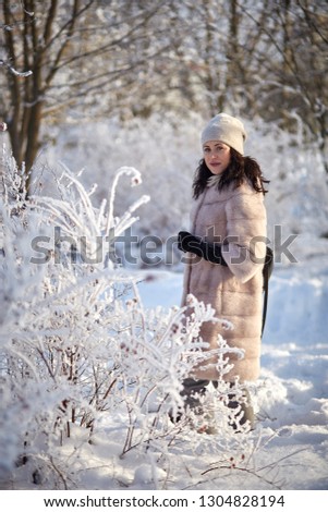 a beautiful young girl in a light mink coat in a black hat and black gloves smiles and walks through the snow-covered forest in winter all the trees are covered with frost