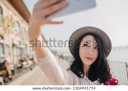 Good-looking white girl in hat using smartphone for selfie. Outdoor portrait of beautiful brunette woman in hat taking picture of herself on sky background.