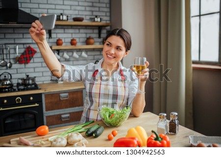 Attractive cheerful woman sit at table in kitchen. She takes selfie and pose on phone camera. Woman hold glass of white wine. Colorful vegetables on table.