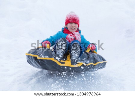 The girl in blue clothes with red hat and scarf descending from the hill covered with a snow on the black rubber ring instead of sledge. The winter picture of holiday recreation