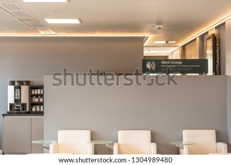 "Silent Lounge" sign (advise) in a empty Lounge area with white chairs, grey wall and coffee maker in the rear 