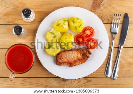 Boiled yellow potatoes with tomatoes, piece of fried carbonnade in white plate, knife, fork, cup of tomato juice, salt, pepper on wooden table. Top view