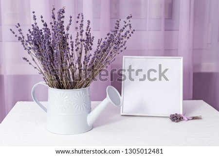Bouquet of dry lavender in watering can with picture frame (mock-up) about window on white table. Soft focus. 