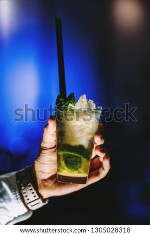 Cold mojito drink on a blue background. Green limes, mint and ice in a glasshouse. Black straw. The man's hand is holding, he gives a drink