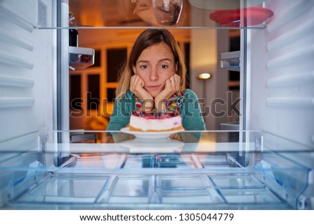 Woman standing in front of fridge with head in hands and looking at cheesecake. Picture taken from the inside of fridge.