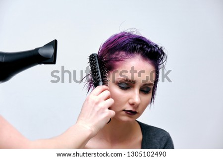 A girl in a gray t-shirt and makeup sitting with wet, purple and short hair on a white background in the middle of the frame. Her hair dries master using a Hairdryer and towels