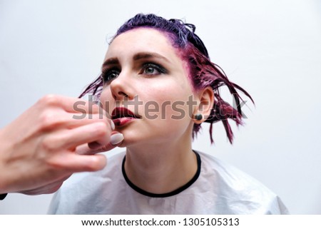 Beautiful girl with violet hair on white background master in make-up. Visible hand of the master and his makeup. The girl is in the middle of the frame and sits on a chair.