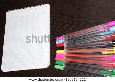 A blank notebook with checkered sheets, a number of multi-colored pens, on a dark wooden background.